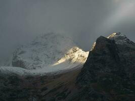 Atmospheric landscape with sharp rocks and high snowy mountain top in blizzard low clouds at overcast. Dramatic gloomy scenery with large snow mountains and glacier in gray cloudy sky at sunrise. photo