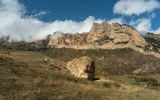 Beautiful autumn mountain landscape with giant granite stone among on hill and mountains. Colorful highland scenery with big rock among fall vegetation and mountains and white clouds on blue sky. photo