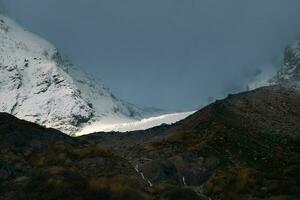 estrecho rayo de luz de sol en un glaciar. contrastando montañas a amanecer. Mañana montaña paisaje con Nevado rock en tormenta de nieve. naturaleza antecedentes de rocoso montaña con agudo rock y nublado cielo. foto