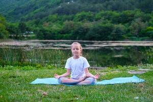 hermosa contento niña con cerrado ojos practicando yoga en el loto posición cerca el lago en naturaleza. niño aprendizaje aptitud física, extensión, yoga, activo estilo de vida conceptos foto