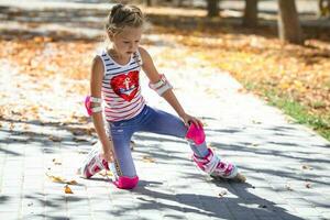 un pequeño niña en rodillo patines y proteccion cayó a el suelo en un otoño parque. niño que cae en el calle con su rodillos foto