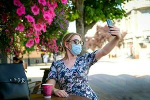 hermosa joven mujer en un café en un Gafas de sol y médico máscara y hace un selfie en el teléfono. un niña en un café en el calle comunica en línea en un teléfono inteligente pandemia COVID-19, cuarentena. foto