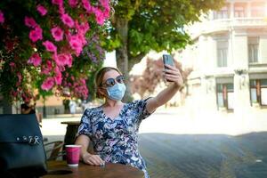 hermosa joven mujer en un café en un Gafas de sol y médico máscara y hace un selfie en el teléfono. un niña en un café en el calle comunica en línea en un teléfono inteligente pandemia COVID-19, cuarentena. foto