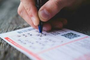 A man is hand crosses out the numbers in a lottery ticket with a pen on a wooden table. Won. Gambling addiction, addiction. photo