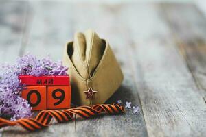 Wooden calendar with Russian text May 9, garrison cap, george ribbon and a bouquet of lilacs on a wooden background. Victory Day. photo