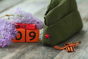 Wooden calendar with Russian text May 9, garrison cap, george ribbon and a bouquet of lilacs on a wooden background. Victory Day. photo