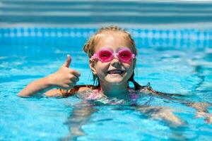 linda pequeño niña sonriente en gafas de protección en el piscina en un soleado día. el niño demostración pulgares arriba me gusta gesto mirando cámara . verano vacaciones foto