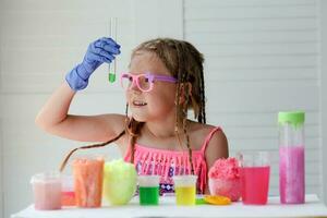 un pequeño niña en rosado lentes conduce experimentos, mira a el cámara y sonrisas un niño en caucho guantes toma un verde líquido con un pipeta y agrega eso a el prueba tubo. experimento. foto