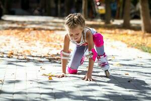 un pequeño niña en rodillo patines y proteccion cayó a el suelo en un otoño parque. foto