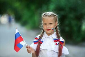 un colegio niño soportes en el al aire libre y sostiene el bandera de Rusia. pequeño niña con cara pintura de ruso simbolismo. ruso bandera día. foto