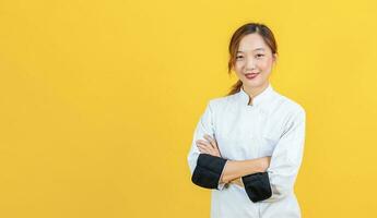 retrato estudio Disparo de asiático mujer cocinero con profesional uniforme posando con cruzar mano en confidente aislado en amarillo antecedentes para culinario y restaurante concepto. foto