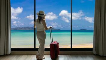 Back view of woman with luggage standing at the window of luxury tropical resort looking at the natural deep blue ocean and white sand beach for summer holiday vacation concept photo