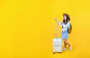 Pretty Asian woman passenger in trendy fashion is pointing while carrying her luggage bag in happiness for travel and summer vacation isolated on yellow background with copy space. photo