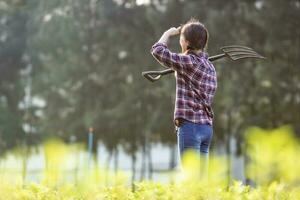 Rear view portrait of Asian woman farmer is carrying garden fork while standing in her countryside vegetables farm during spring season for healthy diet food photo