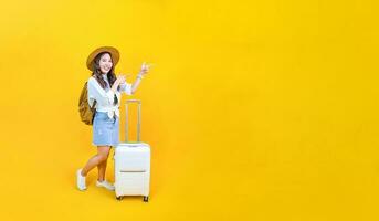 Pretty Asian woman passenger in trendy fashion is pointing while carrying her luggage bag in happiness for travel and summer vacation isolated on yellow background with copy space. photo