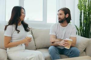 Man and woman sitting at home on the couch in white stylish t-shirts drinking coffee out of crab cups from a coffee shop and having fun chatting smiles and laughter at home. Male and female friendship photo