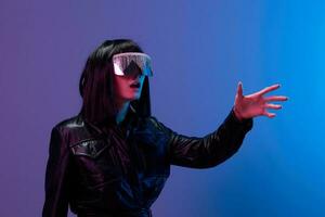 Confused pretty awesome brunet lady in leather jacket trendy specular sunglasses touch invisible object posing isolated in blue violet color light background. Neon party Cyberpunk concept. Copy space photo