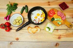 Different food scrambled eggs in frying pan, boiled potatoes, curd, croutons, radishes, cucumbers, tomatoes, smoked sausage, croutons, mint, sorrel on wooden table photo