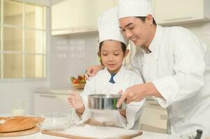 joven asiático padre y su hijo vistiendo cocinero uniforme horneando juntos en cocina a hogar foto