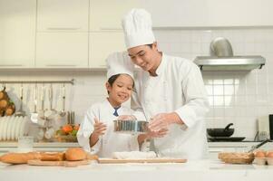 joven asiático padre y su hijo vistiendo cocinero uniforme horneando juntos en cocina a hogar foto