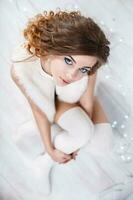 Pretty woman with curls in a fur sleeveless jacket and knitted stockings sitting on the white wooden floor. Top view. photo