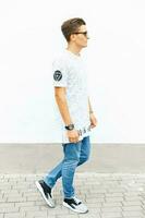 joven hermoso hombre en un blanco camiseta, azul pantalones y zapatillas en pie cerca un blanco pared. foto
