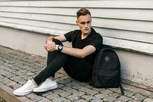 Handsome young man with hairstyle in black fashionable clothes sits near wooden wall and looking sideways photo