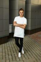 Handsome young hipster stylish man in white shirt, black pants and white shoes posing near modern gray wall. photo