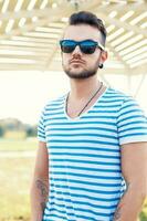 joven hermoso hipster chico con barba en un elegante camiseta y Gafas de sol en un soleado verano día a el playa. foto