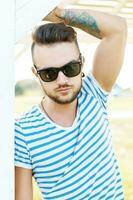 Handsome hipster man in sunglasses with a tattoo on the beach on a sunny day photo