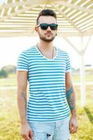 Trendy handsome hipster man with a beard in sunglasses in a trendy shirt stands on the beach. photo