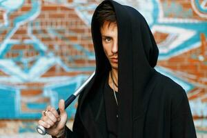 Stylish handsome man in black clothes with a hood standing near a brick wall with graffiti. Man holding bat. photo