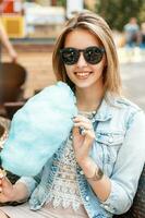 bonito mujer en Gafas de sol y un mezclilla chaqueta comiendo algodón caramelo en el parque. foto