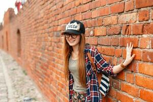 Beautiful cheerful girl hipster smiling in shirt with glasses near a brick wall photo