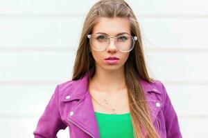 Stylish portrait of a beautiful young woman in a purple jacket with sunglasses near a white wall photo