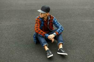 Stylish young man in a black cap, shirt, jeans and sneakers sits on the pavement. photo