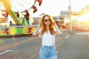 joven hermosa niña con brillante rojo pelo en pie en el diversión parque en un verano día a puesta de sol. foto