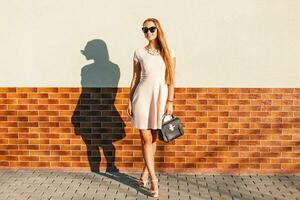Fashionable beautiful woman in sunglasses, pink dress holding bag near brick wall photo