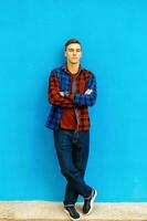 elegante hermoso hombre en un de moda camisa, zapatillas y pantalones soportes cerca un brillante azul pared. foto