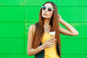 Beautiful young woman with ice cream in sunglasses stands near green wall. photo