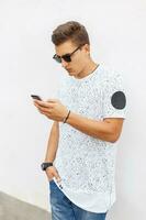 Modern hipster man holding a mobile phone in hands near white wall photo