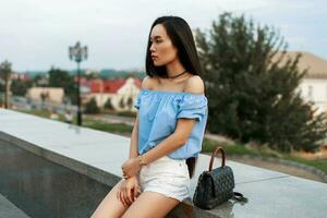 hermosa de moda asiático mujer en un azul blusa y blanco pantalones cortos con un bolso se sienta en un antecedentes de el ciudad. foto