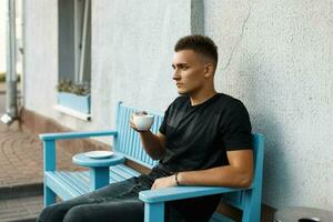 hermoso elegante hombre en un negro camisa sentado en un banco y bebida café foto