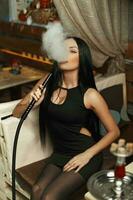 Beautiful girl in stylish black dress smokes a hookah and lets the smoke. photo