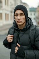 Handsome stylish young man in a black hat and a winter jacket with a backpack in the street photo