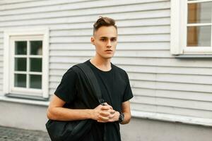 hermoso elegante chico en un negro camiseta con un mochila en pie cerca un de madera casa. foto