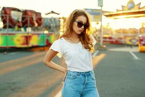hermosa mujer en Gafas de sol y un blanco camiseta en el parque a puesta de sol. foto