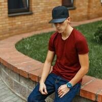 Trendy handsome guy in a black baseball cap sits near a brick building. photo