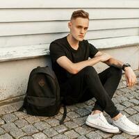 Young handsome guy in a trendy black dress and white shoes sitting near wooden wall. Black backpack. photo