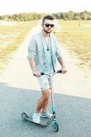 Stylish handsome man in fashionable clothes riding a Kick scooter on the beach photo